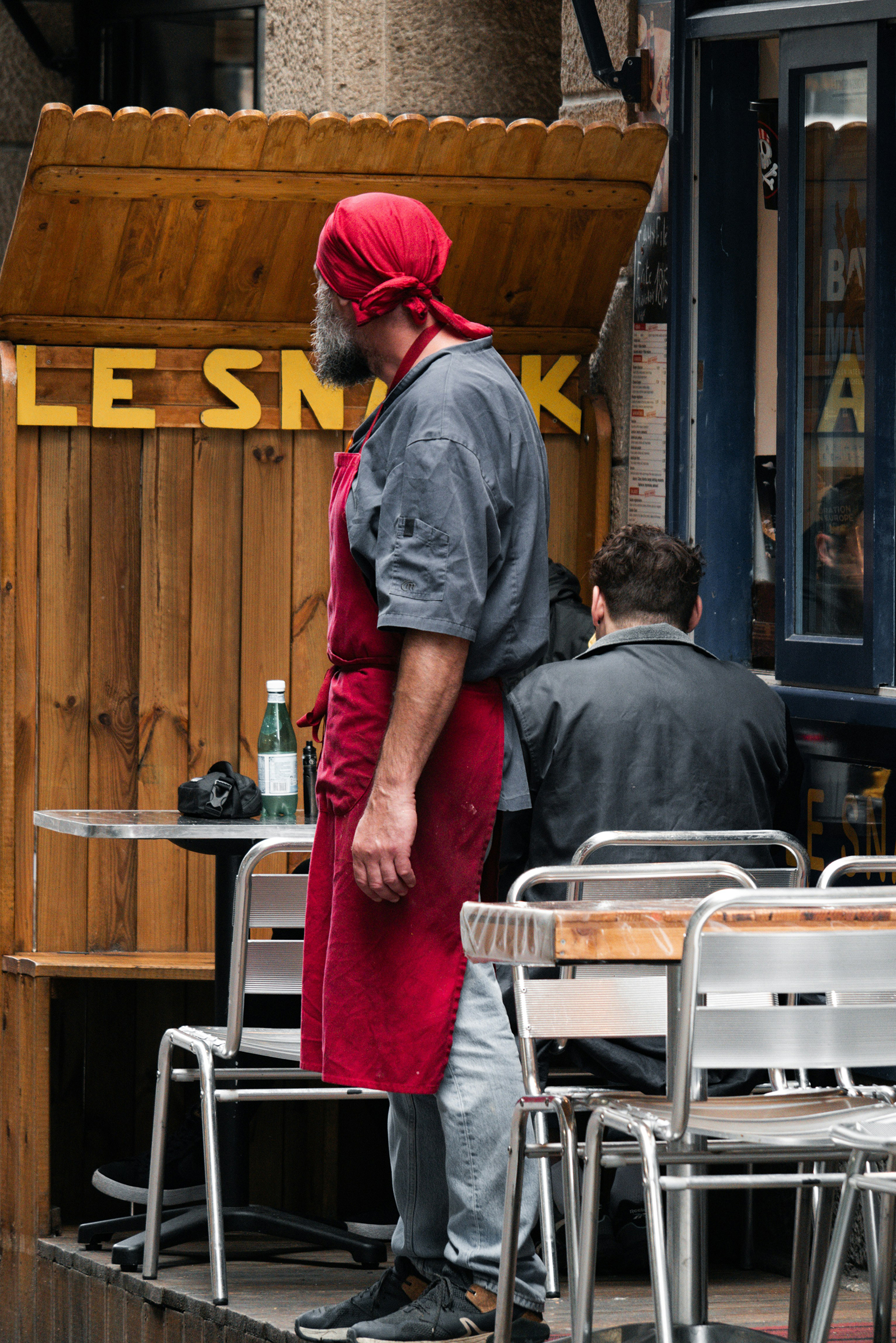 A bearded man in a red apron & bandana stands and looks away in an outdoor cafe